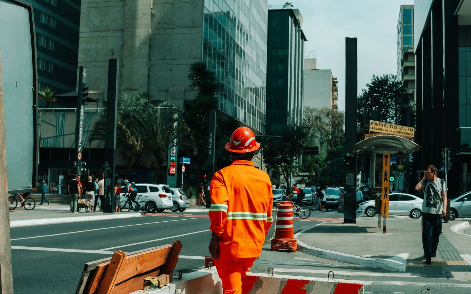 Trabalhador de uniforme laranja atravessa a Avenida Paulista, em São Paulo, entre cones de obra, ciclistas e um ponto de táxi.